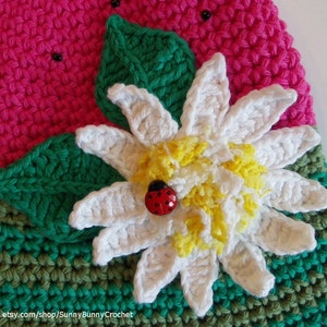 May include: A crocheted watermelon slice with green leaves and a white flower with a yellow center. A ladybug is sitting on the flower.