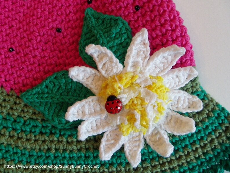 May include: A crocheted watermelon slice with green leaves and a white flower with a yellow center. A ladybug is sitting on the flower.