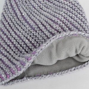 May include: Gray and purple knitted beanie with a fleece lining. The beanie has a diagonal pattern and is decorated with small silver sequins.