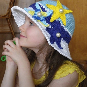 May include: A white crochet sun hat with blue, yellow and purple crocheted flowers and a star. The hat has a wide brim and is decorated with pearl-like beads.
