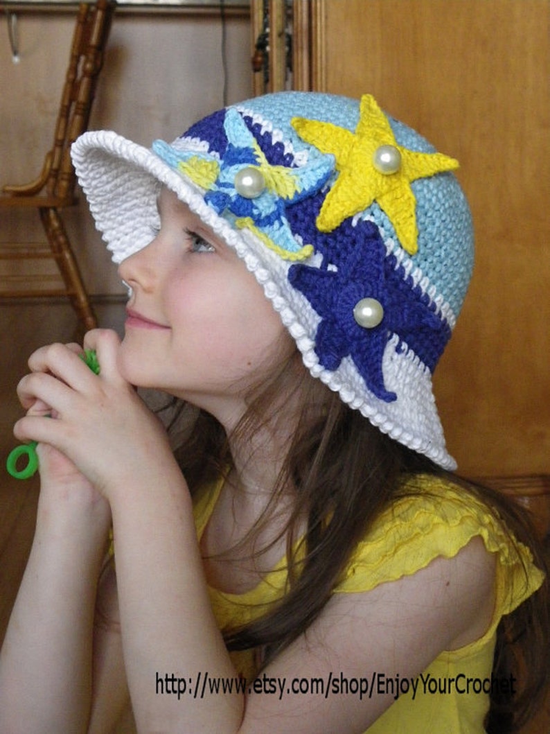 May include: A white crochet sun hat with blue, yellow and purple crocheted flowers and a star. The hat has a wide brim and is decorated with pearl-like beads.