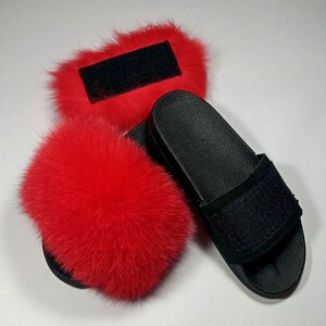 May include: A pair of black faux fur slides with a black strap. The slides are decorated with a red fluffy pom-pom.