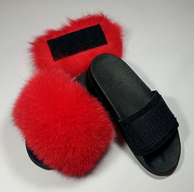 May include: A pair of black faux fur slides with a black strap. The slides are decorated with a red fluffy pom-pom.