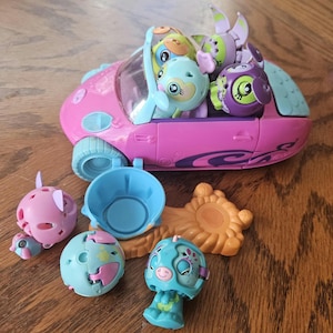 Peut inclure: Une voiture jouet rose avec un intérieur bleu et une variété de personnages jouets ronds et colorés à l'intérieur. La voiture a un design en spirale bleu et violet sur le côté. Il y a aussi plusieurs autres personnages jouets sur la table, dont un personnage bleu et rose, un personnage bleu et blanc et un personnage rose.