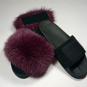 May include: A pair of black leather slides with a burgundy fox fur pom-pom on each strap. The slides have a black velcro strap across the top.