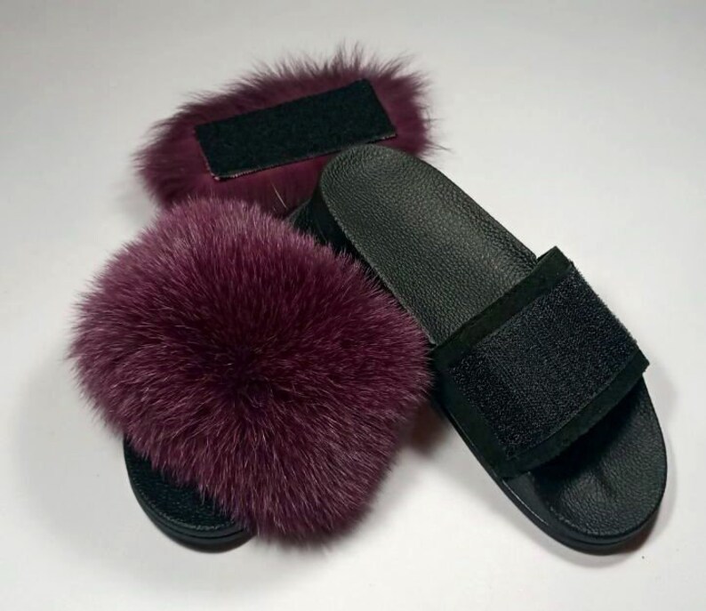 May include: A pair of black leather slides with a burgundy fox fur pom-pom on each strap. The slides have a black velcro strap across the top.