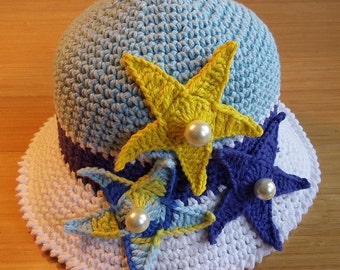 May include: A crochet hat with a white brim and a blue crown. The hat is decorated with three crocheted starfish in yellow, blue, and purple. The starfish have white pearl beads in the center. The hat is labeled "PDF Pattern Only!" and includes a website address.