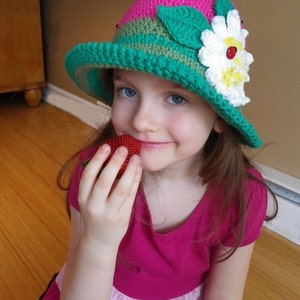 May include: A young girl wearing a pink and green crocheted hat with a white daisy and ladybug embellishment. She is holding a red strawberry in her hand.