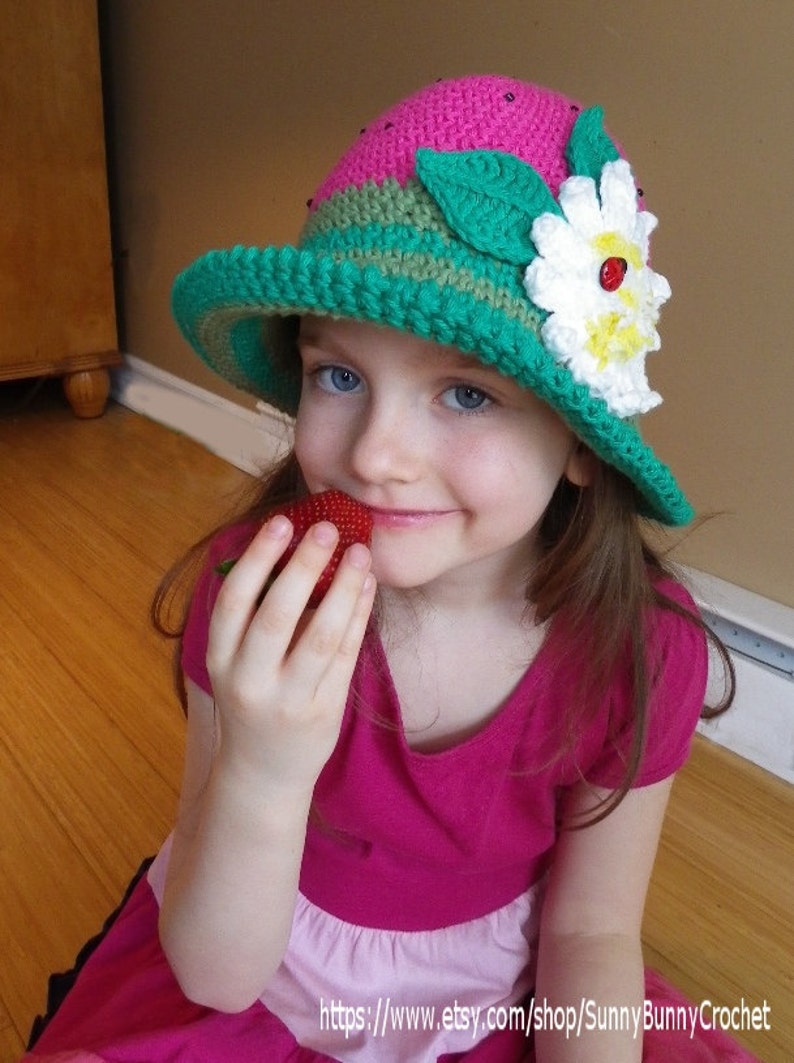 May include: A young girl wearing a pink and green crocheted hat with a white daisy and ladybug embellishment. She is holding a red strawberry in her hand.