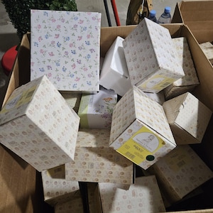 May include: A cardboard box overflowing with boxes of different sizes and designs. Some boxes feature floral patterns in pink, blue, and purple, while others have a repeating baby-themed pattern. The boxes are in various shapes.