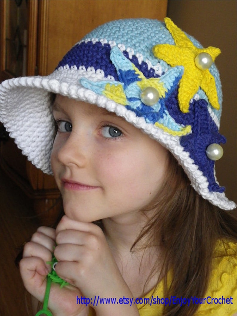May include: A white crocheted sun hat with blue, yellow and white crocheted flowers and pearl accents. The hat is worn by a young girl with long brown hair.