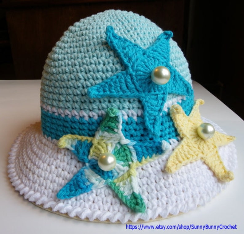 May include: A white crochet sun hat with a wide brim and three crocheted starfish embellishments. The starfish are in shades of blue, green, and yellow, and each has a pearl button in the centre.