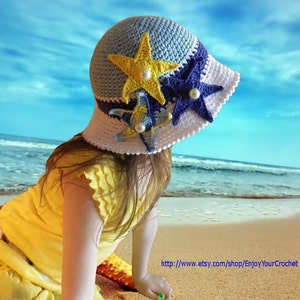May include: A young girl wearing a blue, white and yellow crocheted sun hat with starfish embellishments. The hat has a wide brim and is decorated with three crocheted starfish in different colours. The girl is sitting on a sandy beach with the ocean in the background.