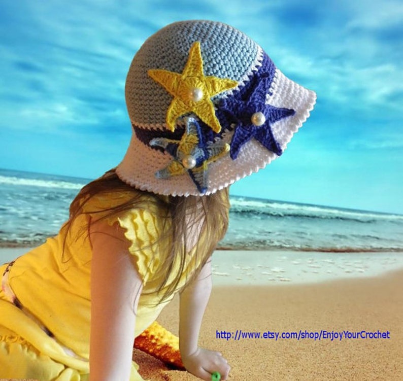 May include: A young girl wearing a blue, white and yellow crocheted sun hat with starfish embellishments. The hat has a wide brim and is decorated with three crocheted starfish in different colours. The girl is sitting on a sandy beach with the ocean in the background.