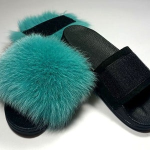May include: A pair of black leather slides with a teal fur pom-pom on the top strap.