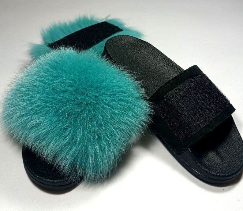 May include: A pair of black leather slides with a teal fur pom-pom on the top strap.