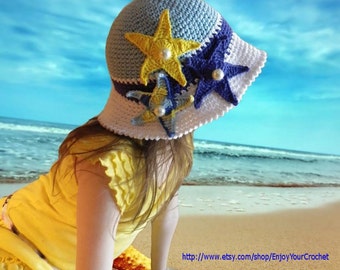 May include: A young girl wearing a white, blue, and yellow crocheted sun hat with starfish embellishments. The hat has a wide brim and is decorated with three starfish, two of which are blue and one is yellow. The girl is standing on a beach with the ocean in the background.