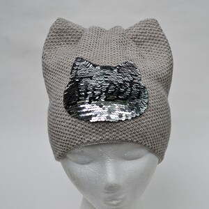 以下が含まれることがあります： グレーのニットビーニーに猫の耳と銀色のスパンコールでできた猫の顔のデザインが施されています。