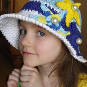 May include: A white crocheted sun hat with blue, yellow, and white crocheted flowers and starfish embellishments. The hat has a wide brim and is decorated with pearl-like beads.