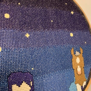 May include: A cross-stitch embroidery hoop with a night sky scene. The sky is a gradient of dark blue to light blue with white stars. There are two figures in the foreground, one purple and one brown.
