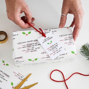 May include: White gift wrap with a red and green holly pattern. The wrapping paper features a humorous guide for cats on how to "destroy" a Christmas tree. The guide includes steps like "destroy the tree", "watch humans put tree back up", "eat", "take a nap", and "see step 1".