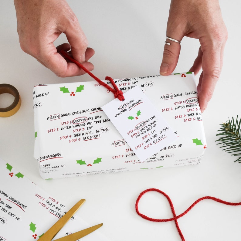 May include: White gift wrap with a red and green holly pattern. The wrapping paper features a humorous guide for cats on how to "destroy" a Christmas tree. The guide includes steps like "destroy the tree", "watch humans put tree back up", "eat", "take a nap", and "see step 1".