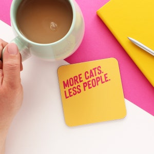 以下が含まれることがあります： ピンクの文字で「MORE CATS. LESS PEOPLE.」と書かれた黄色のコースター