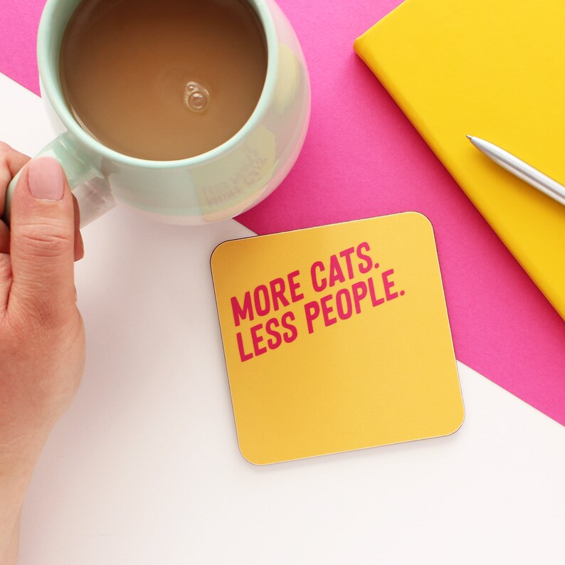 以下が含まれることがあります： ピンクの文字で「MORE CATS. LESS PEOPLE.」と書かれた黄色のコースター
