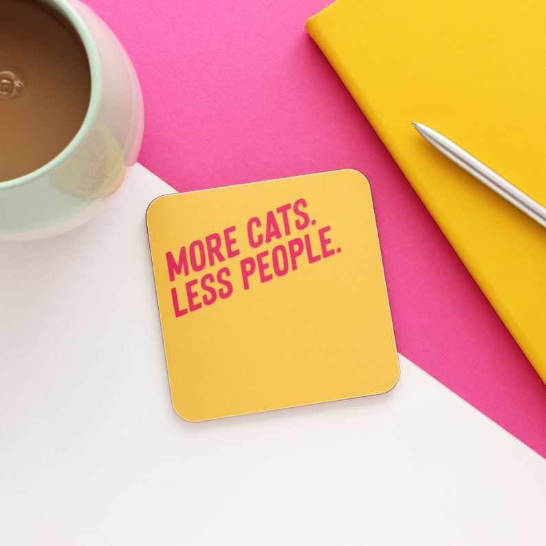 以下が含まれることがあります： ピンクの文字で「MORE CATS. LESS PEOPLE.」と書かれた黄色のコースターです。