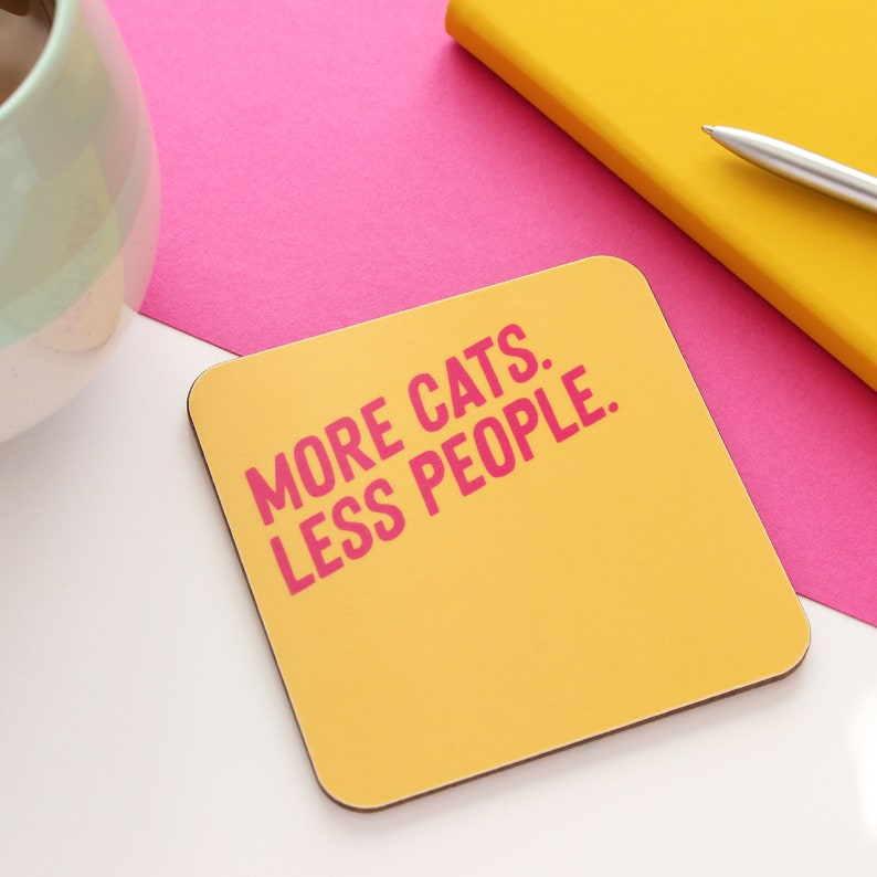 以下が含まれることがあります： ピンクの文字で「MORE CATS. LESS PEOPLE.」と書かれた黄色のコースター