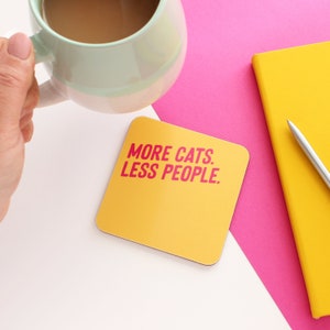 以下が含まれることがあります： ピンクの文字で「MORE CATS. LESS PEOPLE.」と書かれた黄色の正方形コースター