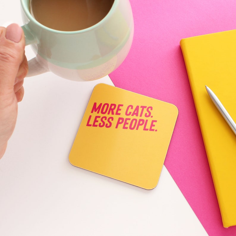 以下が含まれることがあります： ピンクの文字で「MORE CATS. LESS PEOPLE.」と書かれた黄色の正方形コースター
