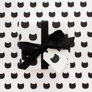 A gift wrapped in cat wrapping paper with a gift tag and decorative black ribbon sitting on a sheet of the wrapping paper.