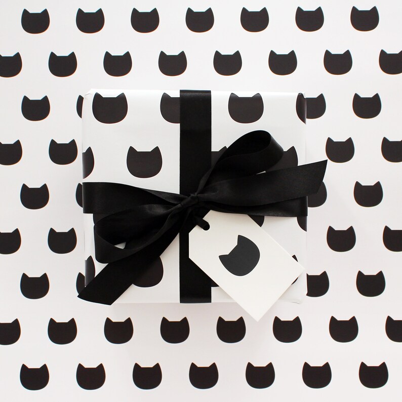 A gift wrapped in cat wrapping paper with a gift tag and decorative black ribbon sitting on a sheet of the wrapping paper.