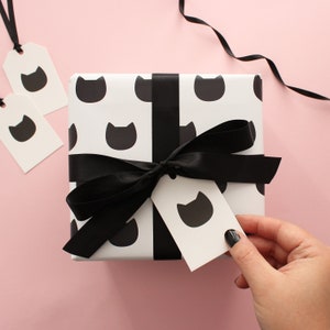A hand holding a traditional gift tag with a single black cat icon attached to a wrapped gift.