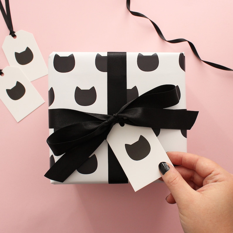 A hand holding a traditional gift tag with a single black cat icon attached to a wrapped gift.