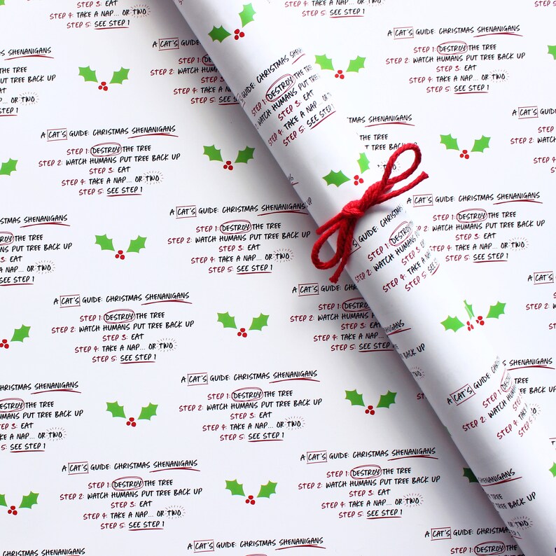 May include: White wrapping paper with a repeating pattern of green holly leaves and red berries. The paper features a festive message that reads "A Cat's Guide to Christmas Shenanigans" with a list of steps: "1. Destroy the tree, 2. Watch humans put tree back up, 3. Eat, 4. Take a nap, or two, 5. See step 1."