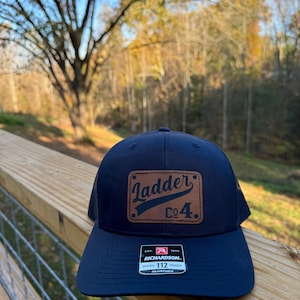 May include: A black trucker hat with a brown leather patch that says "Ladder Co. 4." The hat is on a wooden surface with a metal fence in the background.
