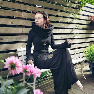May include: A woman wearing a black hooded dress with a long skirt and a pendant necklace. She is sitting on a white bench in front of a wooden fence. There are flowers in the foreground.