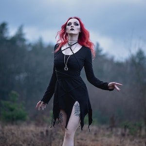 Könnte beinhalten: Eine Frau mit langen roten Haaren trägt ein schwarzes Kleid mit einem Kreuzmuster und eine silberne Halskette. Sie steht auf einem Feld mit einem Wald im Hintergrund.