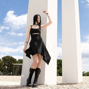 Puede incluir: Una persona con un vestido negro y botas negras se encuentra frente a una estructura de hormigón blanco. La estructura está formada por dos pilares altos y rectangulares con un pilar rectangular más pequeño que los conecta en la parte superior.