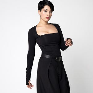 May include: Black long-sleeved top with a square neckline and matching wide-leg trousers. The outfit is accessorised with a black belt and chunky black boots. The model has short black hair.