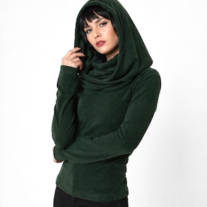 May include: A dark green hooded top with long sleeves and a cowl neck. The hood is draped over the shoulders. The model is wearing black jeans. The fabric looks like a soft, velvet-like material. The top is a deep forest green.