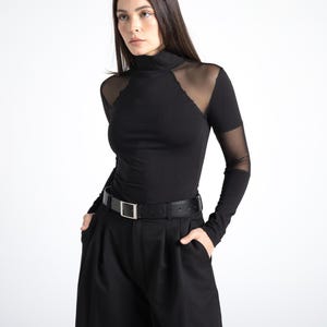 May include: Black long-sleeved top with a high neckline and sheer mesh shoulder and sleeve accents. The top is paired with black wide-leg pants and a black belt with a silver buckle. The outfit is a monochromatic ensemble.