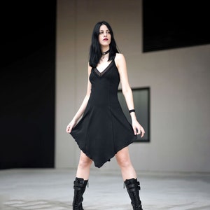 May include: A woman wearing a black dress and black boots. She is standing in front of a white wall.
