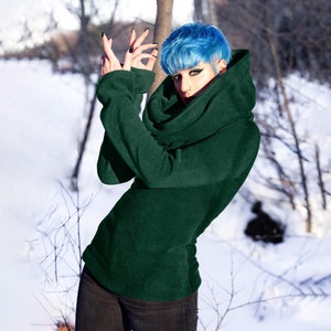 May include: A woman with blue hair is wearing a dark green turtleneck sweater with a large cowl neck. She is standing in a snowy landscape.