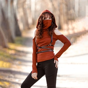 Puede incluir: Una mujer con una camiseta roja con capucha, un arnés de cuero negro y pantalones negros. La camiseta tiene los hombros descubiertos y la capucha está puesta sobre su cabeza. Lleva una máscara facial negra.