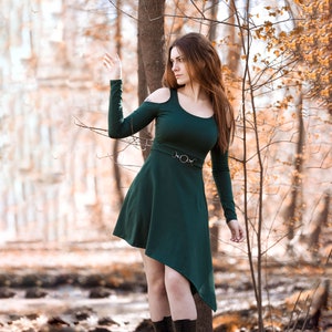 Puede incluir: Una mujer con un vestido verde y un cinturón plateado se encuentra en un bosque con follaje otoñal. El vestido tiene un diseño de hombros descubiertos y un escote alto.