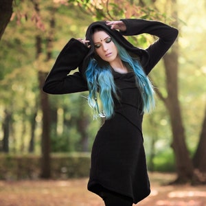 May include: A woman with blue hair wearing a black hooded sweater with a long, flowing design. She is standing in a forest with trees in the background.