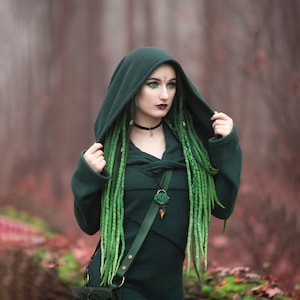 Peut inclure: Une femme portant une cape &agrave; capuche vert fonc&eacute; avec une longue tresse verte. Elle porte un collier avec un pendentif vert et une sangle en cuir. L'arri&egrave;re-plan est une for&ecirc;t floue.